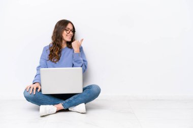 Yerde dizüstü bilgisayarı olan, beyaz arka planda izole edilmiş beyaz bir kadın bir ürünü sunmak için yan tarafı işaret ediyor.