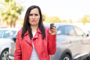 Orta yaşlı kadın dışarıda arabanın anahtarlarını tutarken üzgün bir ifade takınıyor.