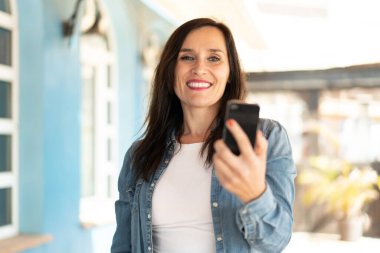 Orta yaşlı bir kadın açık havada mutlu bir ifadeyle cep telefonu kullanıyor.