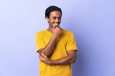 Örgülü genç Afrikalı Amerikalı adam, mor arka planda izole edilmiş, yan tarafa bakıyor ve gülümsüyor.