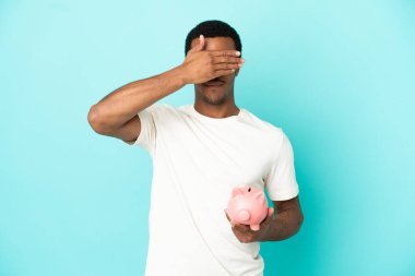 İzole edilmiş mavi arka plan üzerinde kumbarası olan Afro-Amerikalı yakışıklı bir adam elleriyle gözlerini kapatıyor. Bir şey görmek istemiyorum.