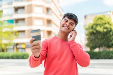 Dışarda cep telefonu kullanan ve elinde mutlu bir ifadeyle kahve tutan genç bir İspanyol.