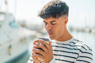 Dışardaki Latin kökenli genç adam elinde kahve tutuyor.