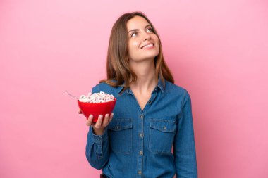 Genç beyaz kadın elinde bir kase mısır gevreği tutarken pembe arka planda izole edilmiş gülümserken yukarı bakıyordu.