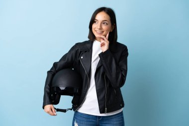 Mavi arka planda motosiklet kaskı tutan bir kadın yukarı bakarken bir fikir düşünüyor.