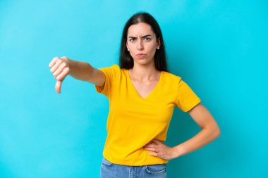 Mavi arka planda izole edilmiş genç beyaz kadın olumsuz ifadeyle baş parmağını aşağı gösteriyor.