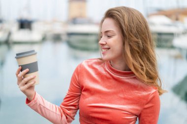 Genç kızıl saçlı kadın açık havada elinde kahveyle neşeli bir ifade ile bekliyor.