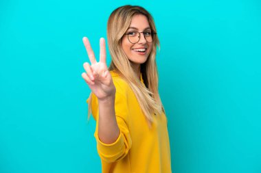 Mavi arka planda izole edilmiş genç Uruguaylı kadın gülümsüyor ve zafer işareti gösteriyor.