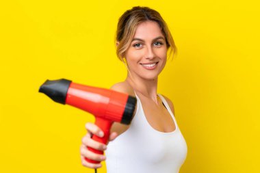 Young Uruguayan woman holding a hairdryer isolate don yellow background with happy expression