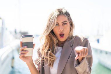 Güzel sarışın Uruguaylı kadın açık havada kahve tutuyor şaşırmış ve ön tarafı işaret ediyor.