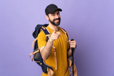 Sırt çantası ve yürüyüş direkleri olan beyaz, yakışıklı bir adam izole arka planda mutlu bir ifadeyle ön tarafı işaret ediyor.