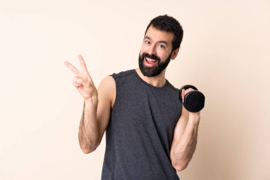 Beyaz sakallı sporcu arka planda halter kaldırıyor. Gülümsüyor ve zafer işareti gösteriyor.