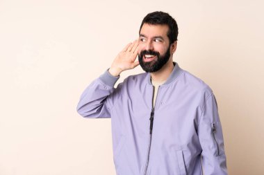 Sakallı, ceketli, soyutlanmış arka planda bir şey dinleyen, elini kulağına koyan beyaz bir adam.