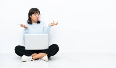 Genç melez kadın dizüstü bilgisayarla yerde oturuyor. Beyaz arka planda, şaşırmış bir yüz ifadesi var.