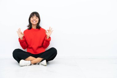 Genç melez kadın, beyaz arka planda izole bir şekilde yerde oturuyor. Parmaklarıyla 