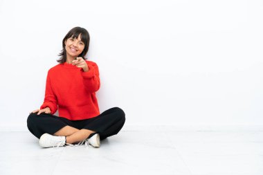 Beyaz arka planda izole bir şekilde yerde oturan genç melez kadın kendinden emin bir ifadeyle sana işaret ediyor.