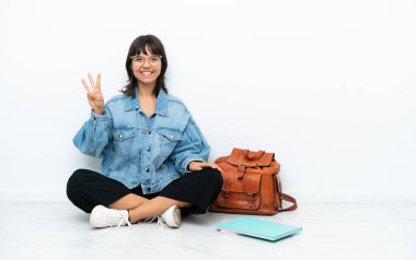 Beyaz arka planda tek bir katta oturan genç bayan mutlu ve parmaklarıyla üçe kadar sayıyor.