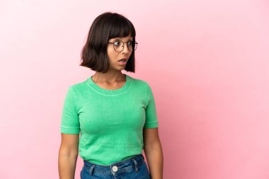 Pembe arka planda soyutlanmış genç melez kadın yan tarafa bakarken sürpriz jest yapıyor.