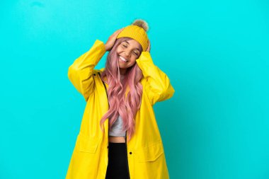 Pembe saçlı, yağmura dayanıklı bir ceket giyen, mavi arka planda izole edilmiş, gülen genç bir kadın.