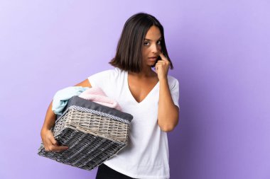 Mor arka planda izole edilmiş çamaşır sepetini tutan genç Latin kadın bir şey gösteriyor.