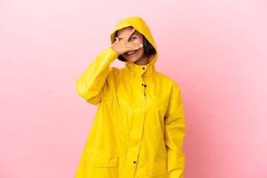 Yağmura dayanıklı bir ceket giyen genç Latin bir kadın izole arka planda gözleri elleriyle kaplıyor ve gülümsüyor.