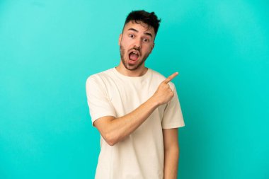 Mavi arka planda izole edilmiş genç beyaz adam şaşırmış ve işaret ediyor.
