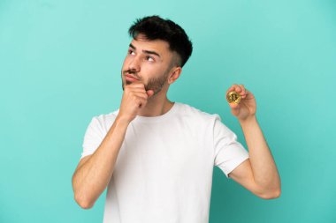 Elinde Bitcoin tutan genç bir adam mavi arka planda izole edilmiş ve yukarı bakıyor