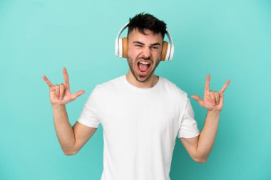 Kafkasyalı genç adam mavi arka planda izole edilmiş müzik dinliyor ve rock hareketleri yapıyor.