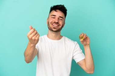 Elinde mavi arka planda izole edilmiş Bitcoin tutan genç adam para hareketi yapıyor.