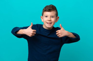 Küçük çocuk mavi arka planda izole edilmiş bir şekilde baş parmak hareketi yapıyor.