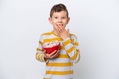 Beyaz arka planda izole edilmiş mısır gevreği kasesi tutan küçük beyaz çocuk mutlu ve ağzı eliyle kaplayan bir gülümseme.