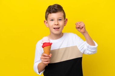 Sarı arka planda izole edilmiş kornet dondurmalı beyaz çocuk zaferi kutluyor.