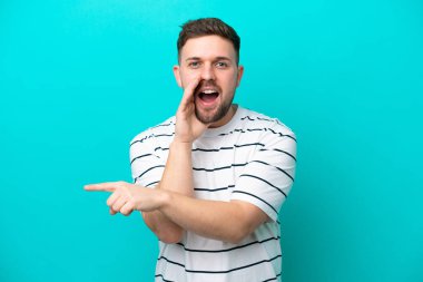 Mavi arka planda izole edilmiş genç beyaz bir adam bir ürün sunmak ve bir şeyler fısıldamak için kenarı işaret ediyor.