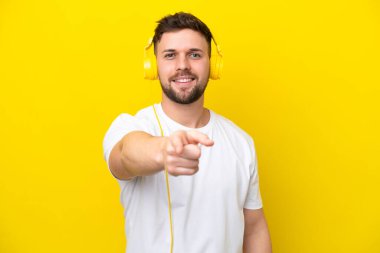 Genç beyaz adam sarı arka planda izole edilmiş müzik dinliyor ve ön tarafı işaret ediyor.