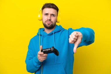 Sarı arka planda izole edilmiş bir bilgisayar oyunu kontrolörüyle oynayan genç beyaz adam, negatif ifade ile baş parmağını aşağı gösteriyor.