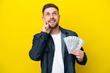 Young caucasian man taking a lot of money  isolated on yellow background thinking an idea while looking up