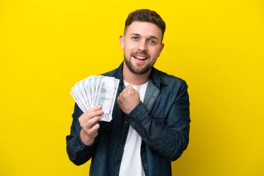 Young caucasian man taking a lot of money  isolated on yellow background celebrating a victory