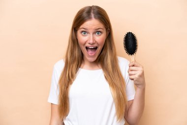 Saçları taranmış genç, sarışın bir kadın bej arka planda, şaşırmış ve şok olmuş bir yüz ifadesi var.