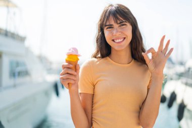Açık havada elinde kornet dondurması olan genç bir kadın parmaklarıyla tamam işareti gösteriyor.