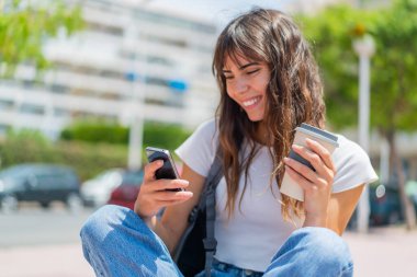 Dışarıda cep telefonu kullanan ve elinde kahve tutan genç bir kadın.