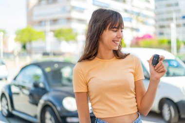 Dışarıda arabanın anahtarlarını tutan genç bir kadın, neşeli bir ifadeyle.