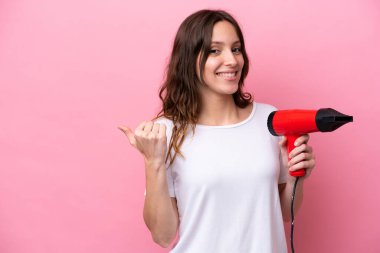 Pembe arka planda izole edilmiş saç kurutma makinesi tutan beyaz bir kadın bir ürünü sunmak için kenarı işaret ediyor.