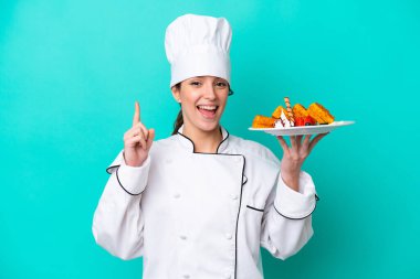 Kafkasyalı genç aşçı kadın elinde waffle tutarken mavi arka planda izole edilmiş harika bir fikre işaret ediyor.