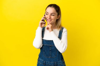 İzole edilmiş sarı arka plan üzerinde cep telefonu kullanan genç bir kadın yana bakıyor ve gülümsüyor.