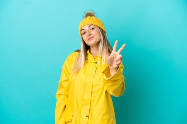 İzole edilmiş mavi arka planda yağmurluk giyen genç bir kadın mutlu ve parmaklarıyla üçe kadar sayıyor.
