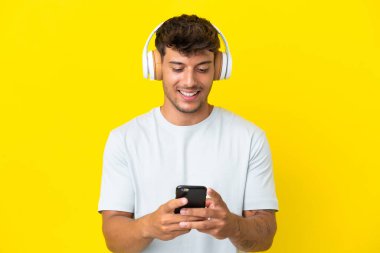 Beyaz tenli yakışıklı adam sarı arka planda izole edilmiş müzik dinliyor ve cep telefonu arıyor.