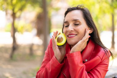 Orta yaşlı bir kadın elinde mutlu bir ifadeyle avokado tutuyor.