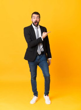 İzole edilmiş sarı arka plan üzerinde tam boy iş adamı görüntüsü. Şaşırmış ve işaret ediyor.