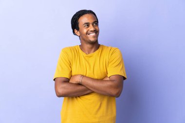 Örgülü genç Afrikalı Amerikalı adam mor arka planda izole edilmiş gülümserken yukarı bakıyor.