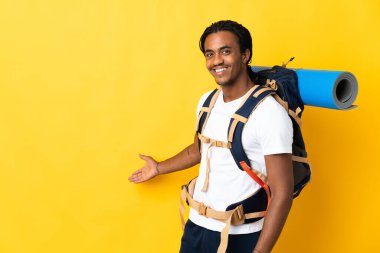 Sarı arka planda izole edilmiş büyük bir sırt çantalı genç dağcı. Davet etmek için ellerini yana uzatıyor.